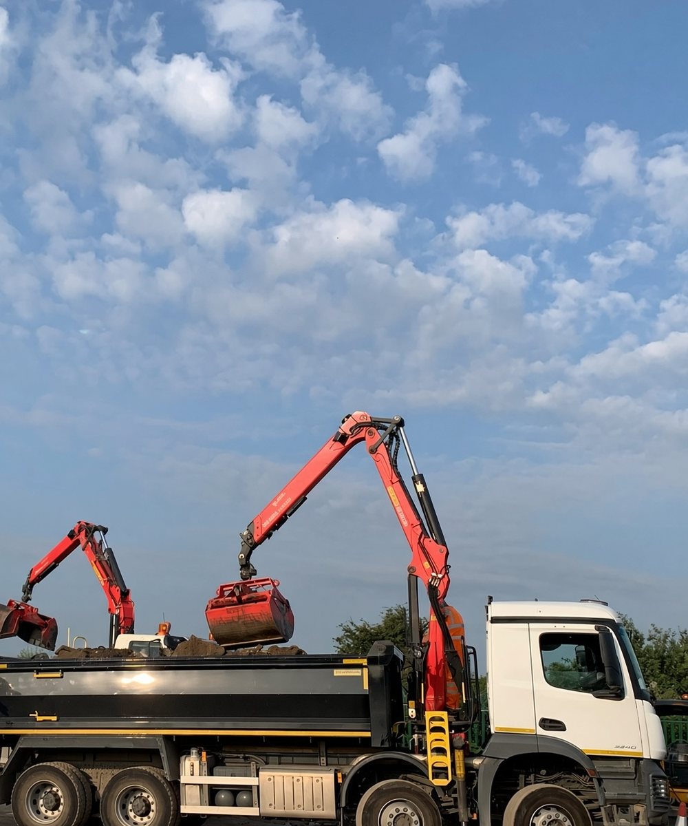 Two SAUK grab lorries loading material, crane arms raised against a blue sky